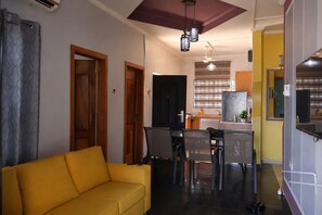 Una sala de estar con un sofá amarillo, una zona de comedor con sillas y una mesa, y una cocina al fondo.