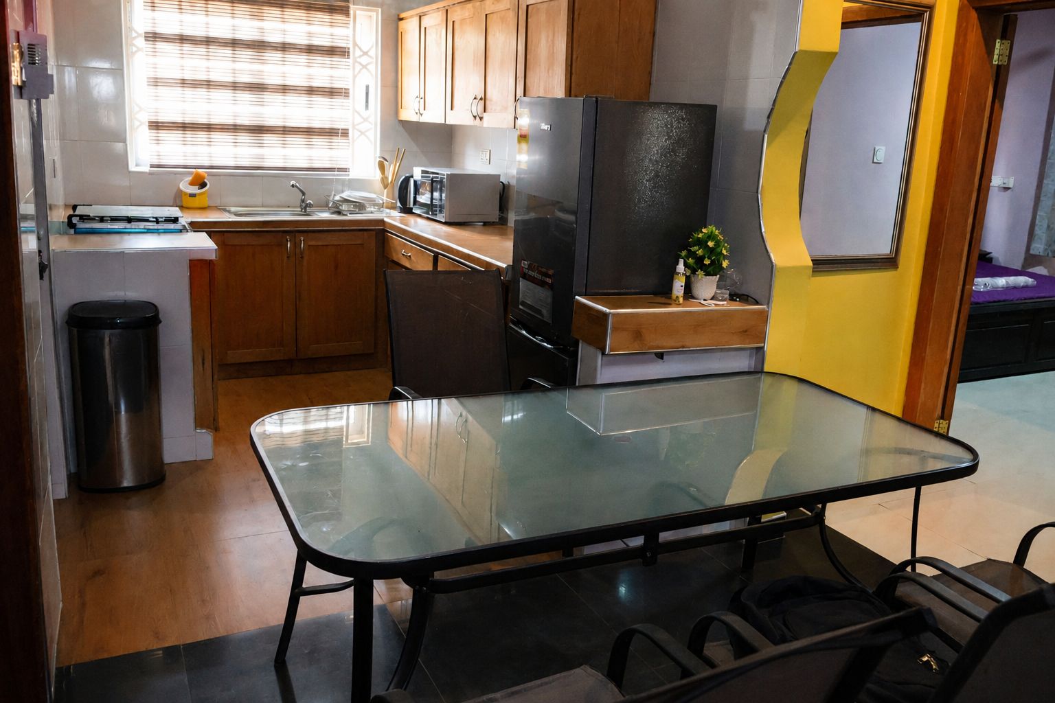 Una cocina compacta con armarios de madera, un cubo de basura de acero inoxidable, una mesa de vidrio y un refrigerador.