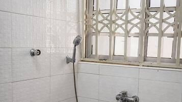 A bathroom with white tiled walls, a shower with a handheld showerhead, a window with a decorative grille, and a towel rack with a towel.