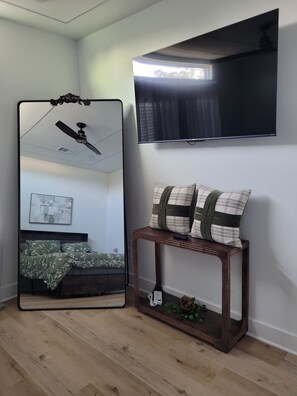 Een kamer met een bed, een spiegel, een aan de muur bevestigde televisie en een klein houten nachtkastje met decoratieve kussens.