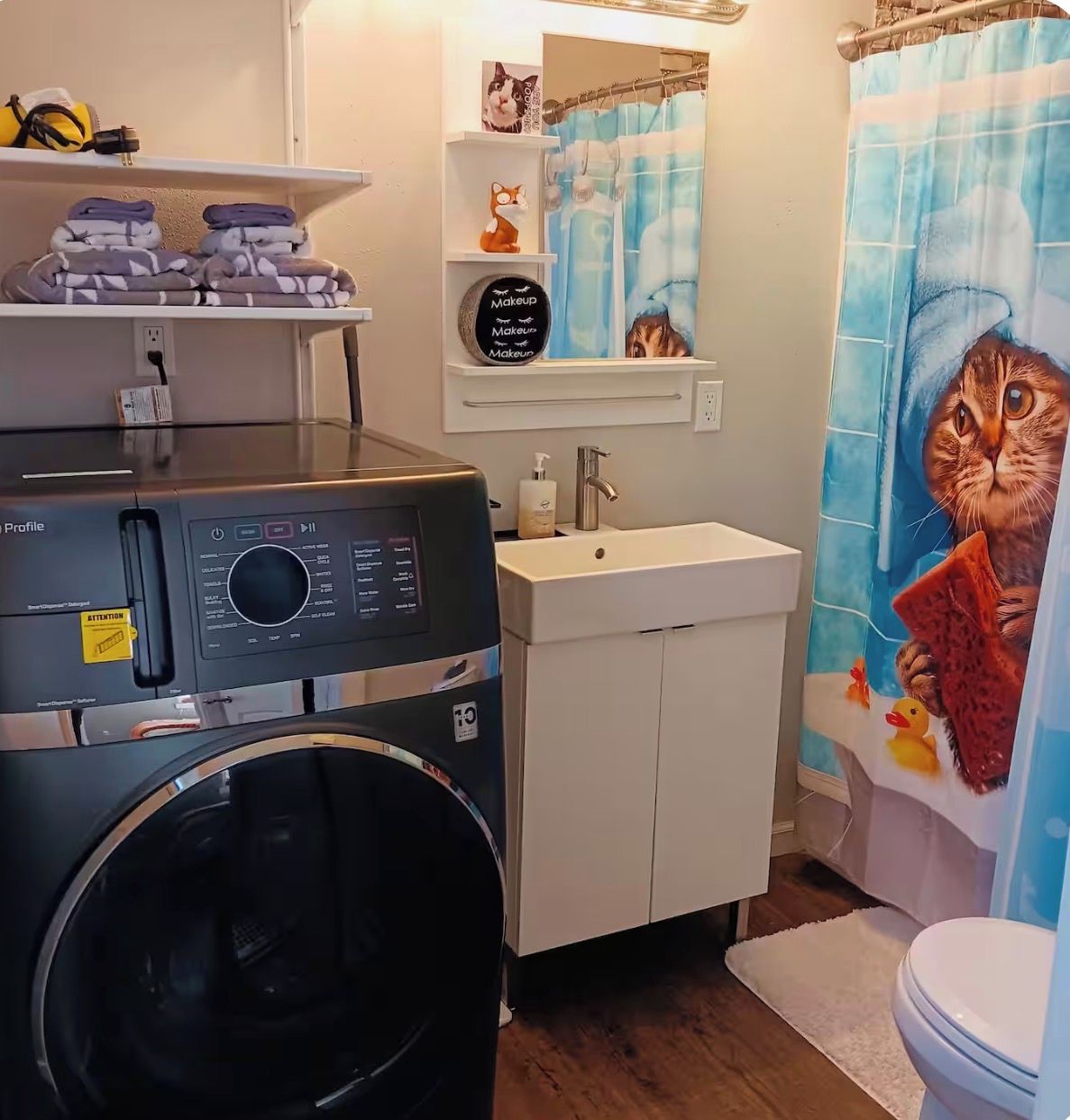 A bathroom with a washing machine, a sink, a mirror, and a shower curtain with a cat design.