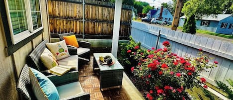 A covered patio with wicker furniture, a small table, and a flower bed with red flowers.