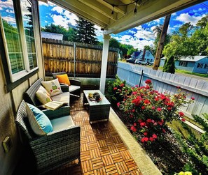 A covered patio with wicker furniture, a small table, and a flower bed with red flowers. - Mr. Fatness Home: A puurrrfet place to relax & make unique memories 🐾🐈🥰 (Kittitas)