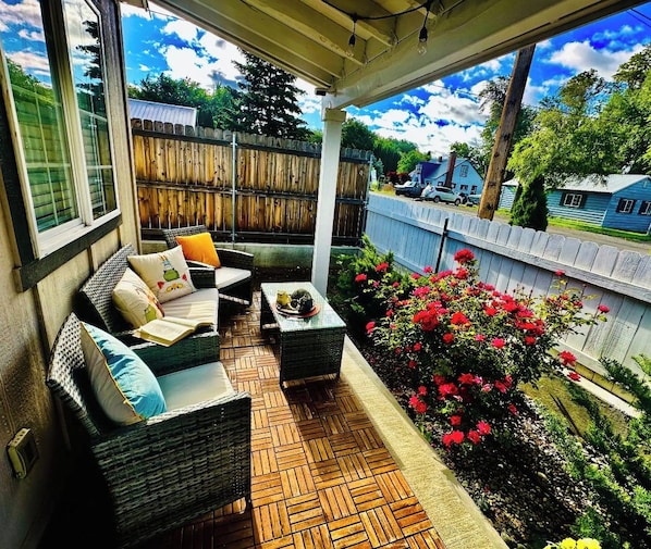 A covered patio with wicker furniture, a small table, and a flower bed with red flowers.