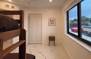 A room with a bunk bed, a small stool, a door with louvered shutters, a framed picture on the wall, and a window with a view of parked cars. - Luxury Miami Stay • Near Wynwood/Design District (Miami)