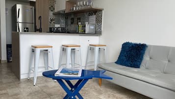 Eine kompakte Küche mit Kühlschrank, Mikrowelle und Kaffeemaschine. Drei weiße Barhocker und ein blauer Tisch mit einer Zeitschrift. Im Wohnbereich befindet sich ein graues Sofa mit einem blauen Sitzkissen.
