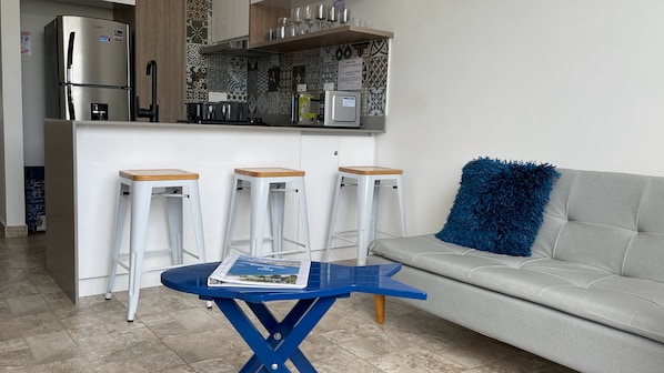 Een kleine keuken met een koelkast, magnetron en koffiezetapparaat. Drie witte barstoelen en een blauwe tafel met een tijdschrift. In de woonkamer staat een grijze bank met een blauw sierkussen.