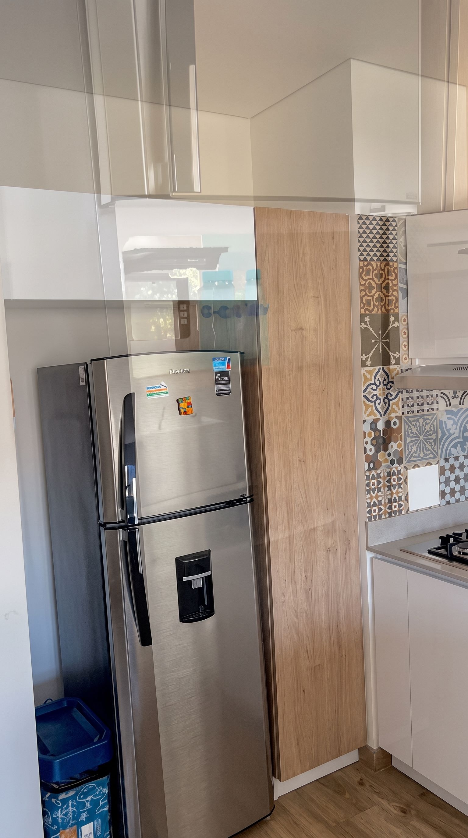 Ein Edelstahl-Kühlschrank mit Wasserspender, neben einem Holzschrank und einer gefliesten Spritzschutzwand.