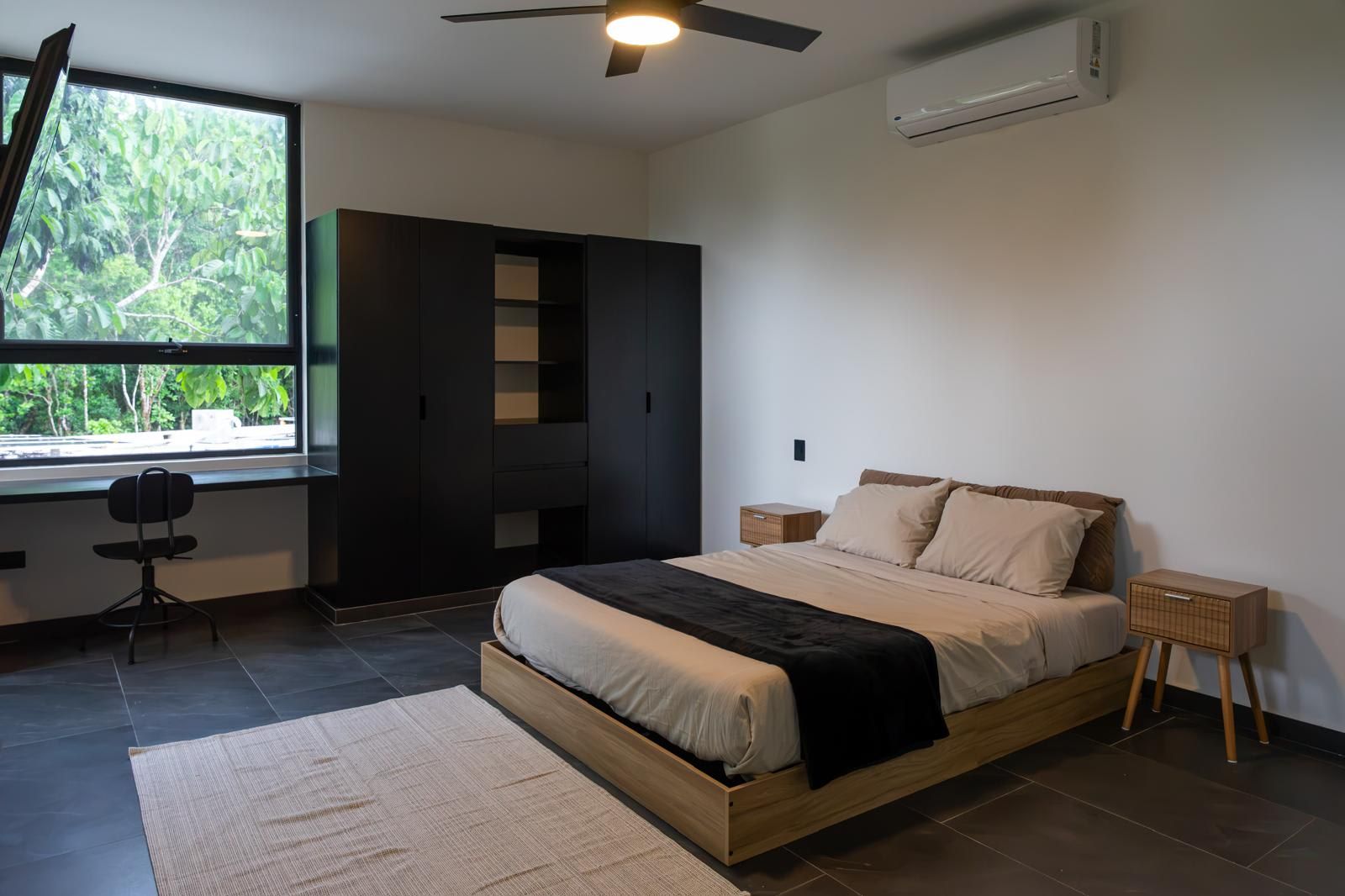 A bedroom with a bed, a desk, a wardrobe, and a window with a view of greenery.