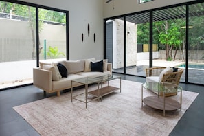 A modern living room with a sofa, coffee table, and chair, large windows, and a view of a swimming pool. - Xpuha Villa, Private Pool and Jungle, Near Beach (Xpu Ha)