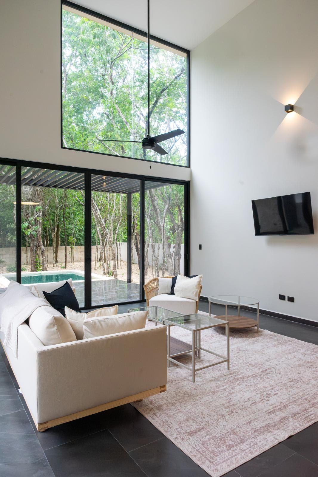A modern living room with a large glass wall, a ceiling fan, a sofa, a coffee table, and a TV mounted on the wall.