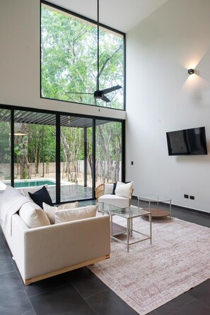 A modern living room with a large glass wall, a ceiling fan, a sofa, a coffee table, and a TV mounted on the wall.