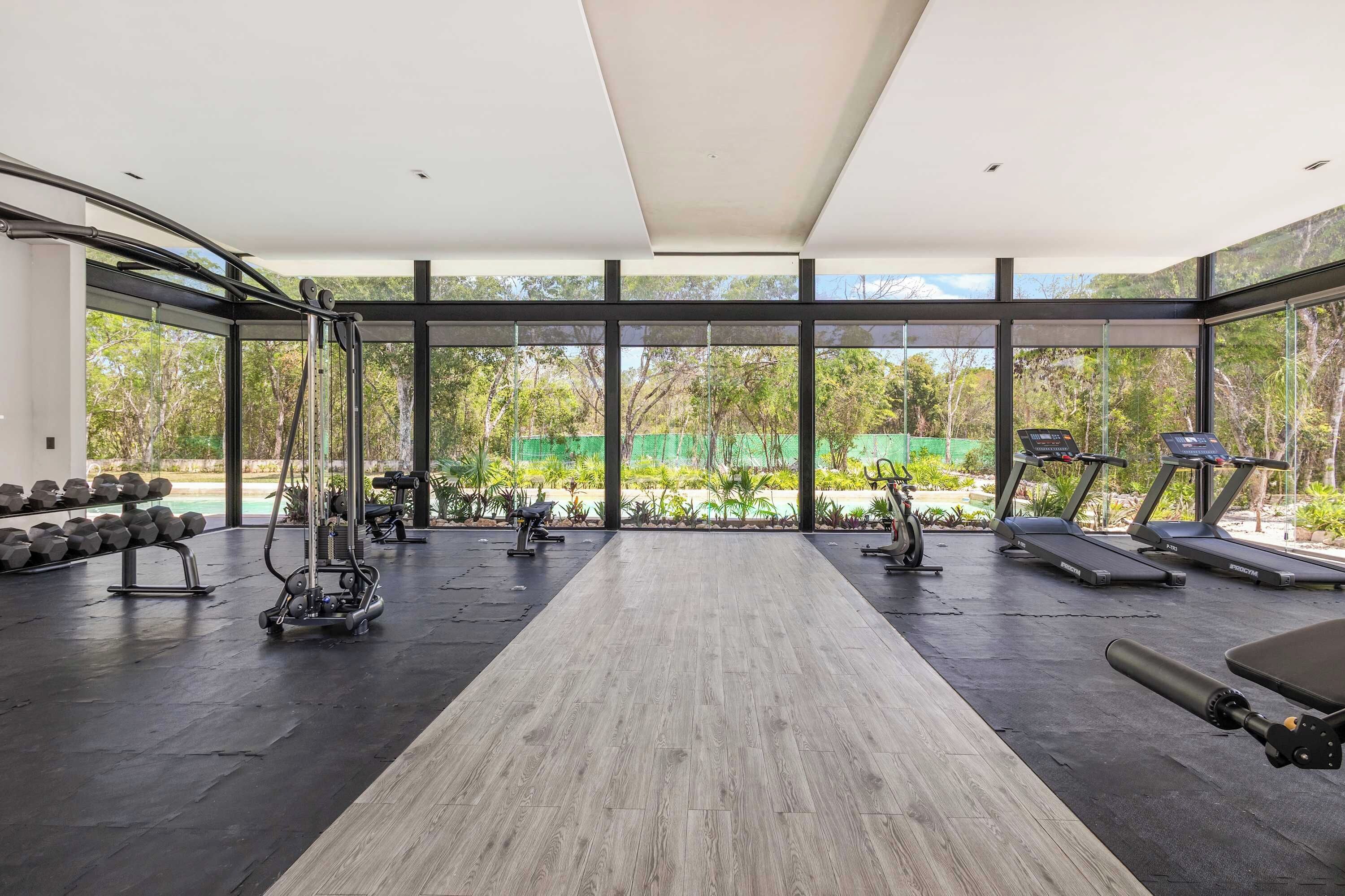 Een sportschool met trainingsapparatuur, een houten vloer en grote ramen met uitzicht op groen.