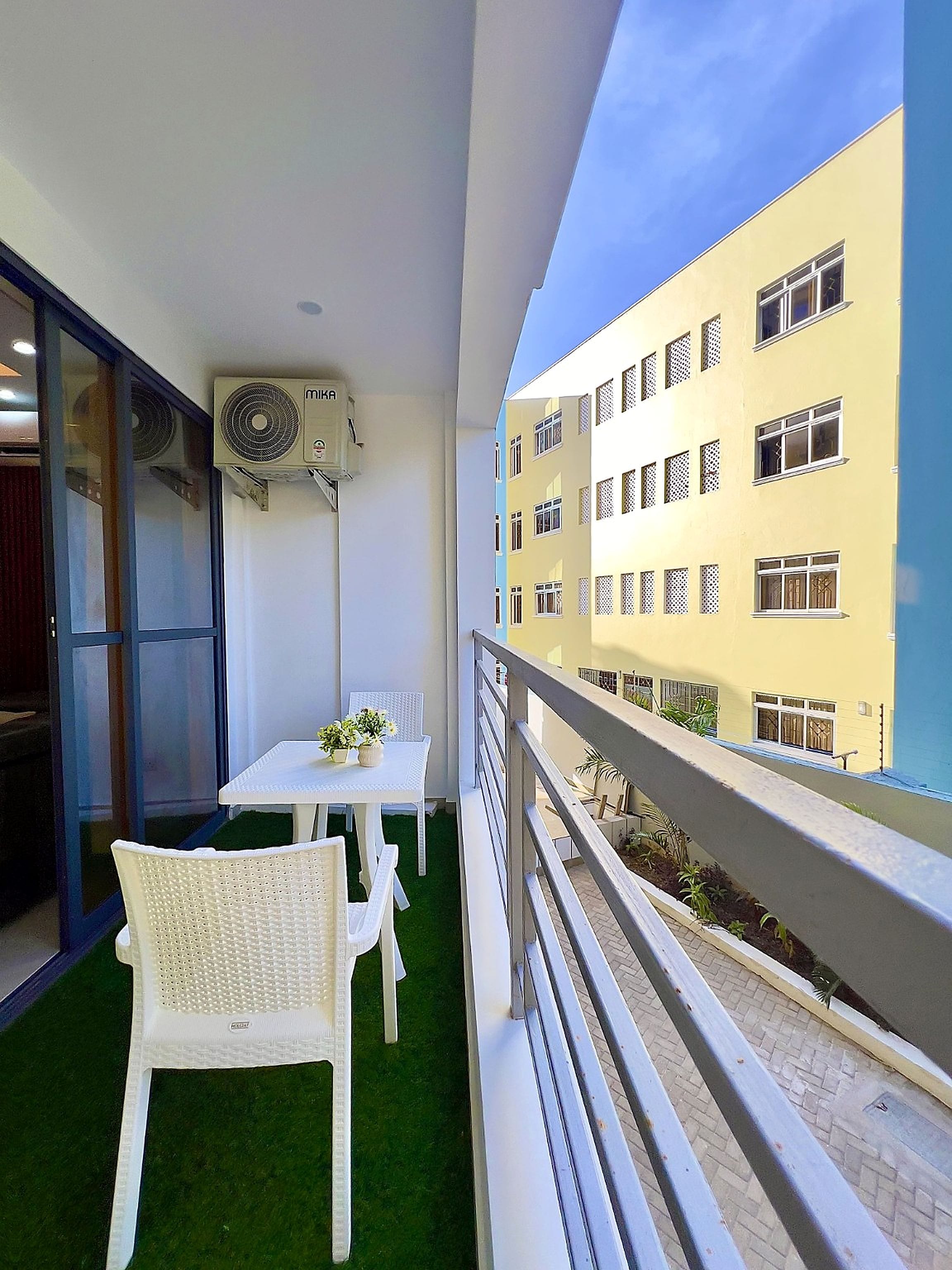 Un balcon avec une table et des chaises, une vue sur un immeuble jaune et un appareil de climatisation.