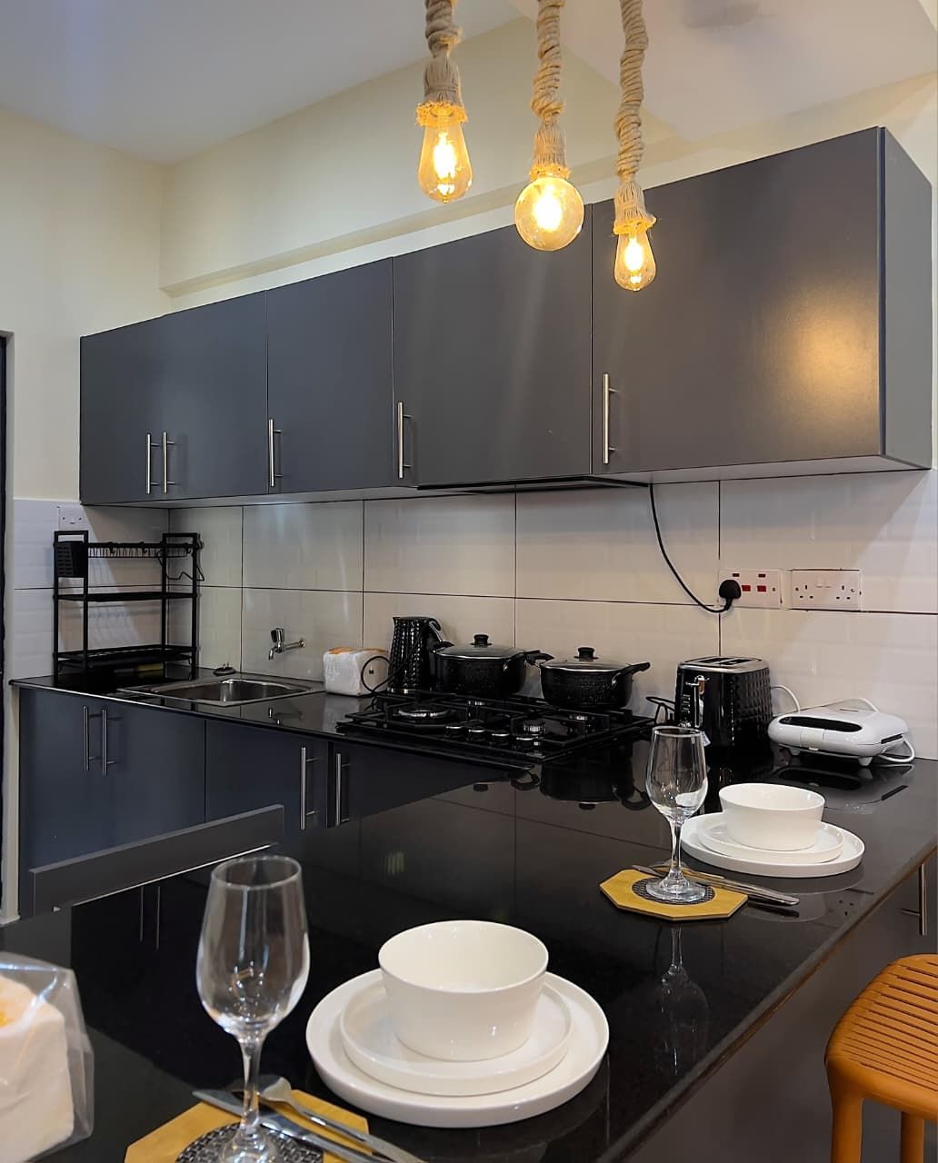 Une cuisine moderne avec des meubles de couleur sombre, un plan de travail noir et un îlot central agrémenté d’assiettes blanches et d’un verre à vin.