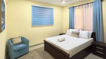 Une chambre à coucher comprenant un lit, une chaise, une table de chevet, un ventilateur de plafond et une fenêtre avec des stores.