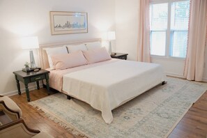 A bedroom with a large bed, two bedside tables, a nightstand, a framed cityscape picture, and a window with curtains. - Child-Friendly Retreat Near Charleston, 2 (Hanahan)