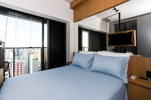 A hotel room with a large bed, a TV mounted on the wall, and a view of the city through the window. - On Paulista 1921 (Vila Mariana)