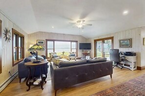 A spacious living room with a large window, a ceiling fan, a sofa, a dining table, and a computer desk. - Lakefront, dog-friendly house with amazing view, dock, 2 kayaks & canoe (Isle La Motte)