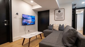 A modern living room with a large flat-screen TV mounted on the wall, a grey sofa with multiple cushions, a small marble-top table, and a dark door.
