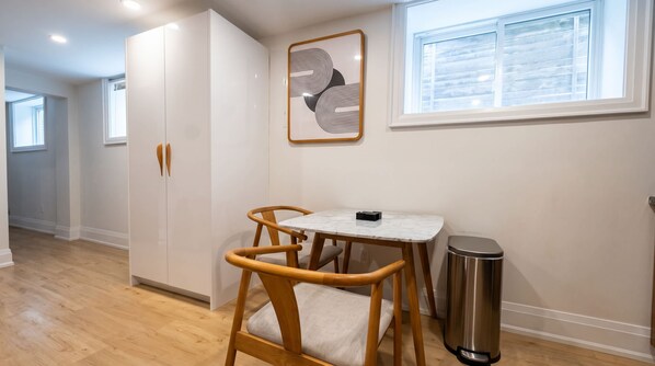 A compact kitchen with a white cabinet, a small table with chairs, a trash bin, and a window with blinds.