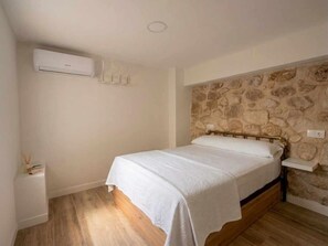 A bedroom with a bed, a wall-mounted air conditioner, a stone-patterned wall, and a wooden floor.