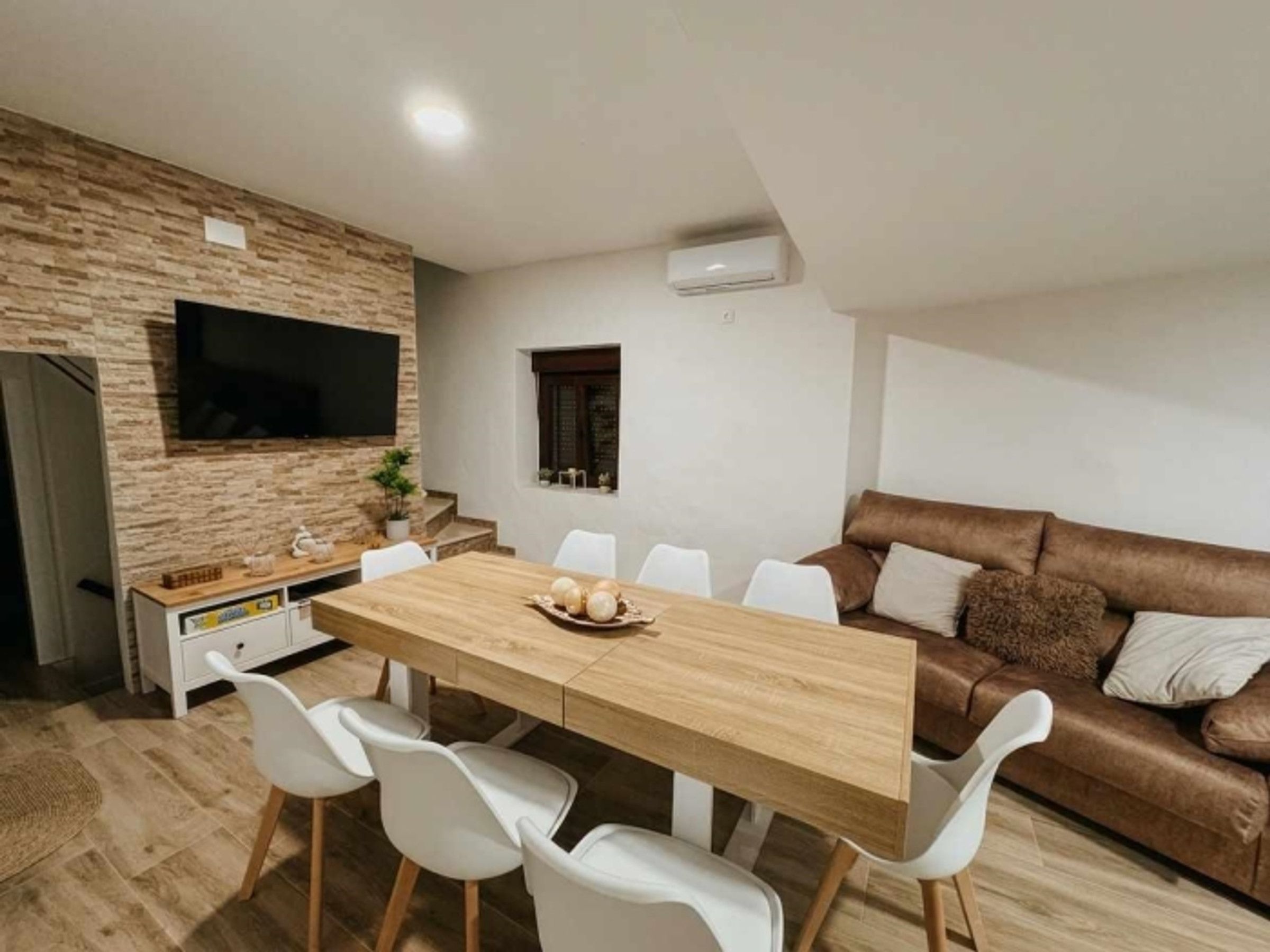 Un salon moderne avec une table à manger en bois, des chaises blanches, un canapé en cuir marron, un téléviseur à écran plat fixé à un mur en pierre et une petite fenêtre.