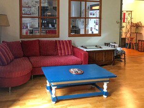 Sala de estar com um sofá vermelho, uma mesa de centro azul e uma área de cozinha visível pela janela.