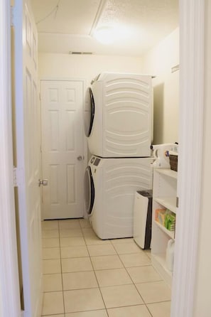 Un lavadero con lavadoras y secadoras apiladas, una puerta y estantes con productos de limpieza.