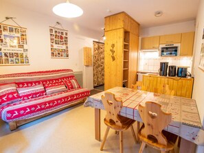 A cozy living room with a red patterned sofa, wooden dining table with chairs, and a kitchen area with wooden cabinets and appliances. - Quiet apartment with terrace - 4 guests (Villarodin-Bourget)