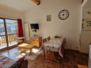 A room with a wooden floor, a dining table with a tablecloth, a television on a stand, a clock on the wall, and a window with a view of buildings. - 3-room apartment, south balcony - 6 guests (Modane)
