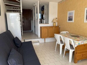 A compact living space with a sofa, a dining table with chairs, a refrigerator, a wooden cabinet, and a kitchen area with a window. - Duplex with parking - 6 guests in quiet area (Le Grau-du-Roi)