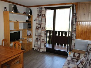 A room with a wooden table, a television on a shelf, a sofa, and a sliding glass door leading to a balcony. - Quiet apartment, forest - 4 guests (Villarodin-Bourget)
