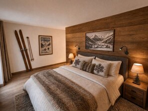 Un dormitorio con cabecera de madera, cama con una manta de piel, dos almohadas con diseños de copo de nieve, un cuadro enmarcado de un paisaje montañoso y un par de esquíes apoyados en la pared.