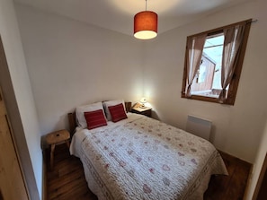A bedroom with a bed, a nightstand, a lamp, a window with curtains, and a hanging light fixture. - Cozy mountain apartment - 4 guests (Arêches-Beaufort)