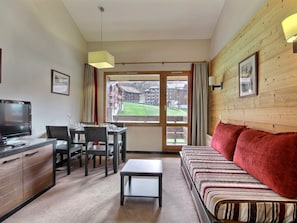 A hotel room with a television, dining table, chairs, a sofa, a small table, and a large window with a view of buildings and a lawn. - Charming duplex, slopes view - 4 guests (Belle Plagne)