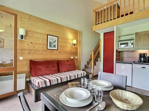 A cozy room with a wooden wall, a red door, a dining table set with plates, glasses, and a spoon, and a kitchen area with a microwave and coffee maker. - Charming duplex, slopes view - 4 guests (Belle Plagne)
