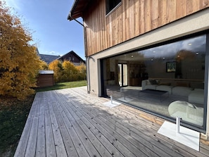 A modern house with a wooden exterior, a deck, and a large glass door leading to a living area with a sofa and a coffee table. - Modern house in village center - 12 guests (Villard-de-Lans)