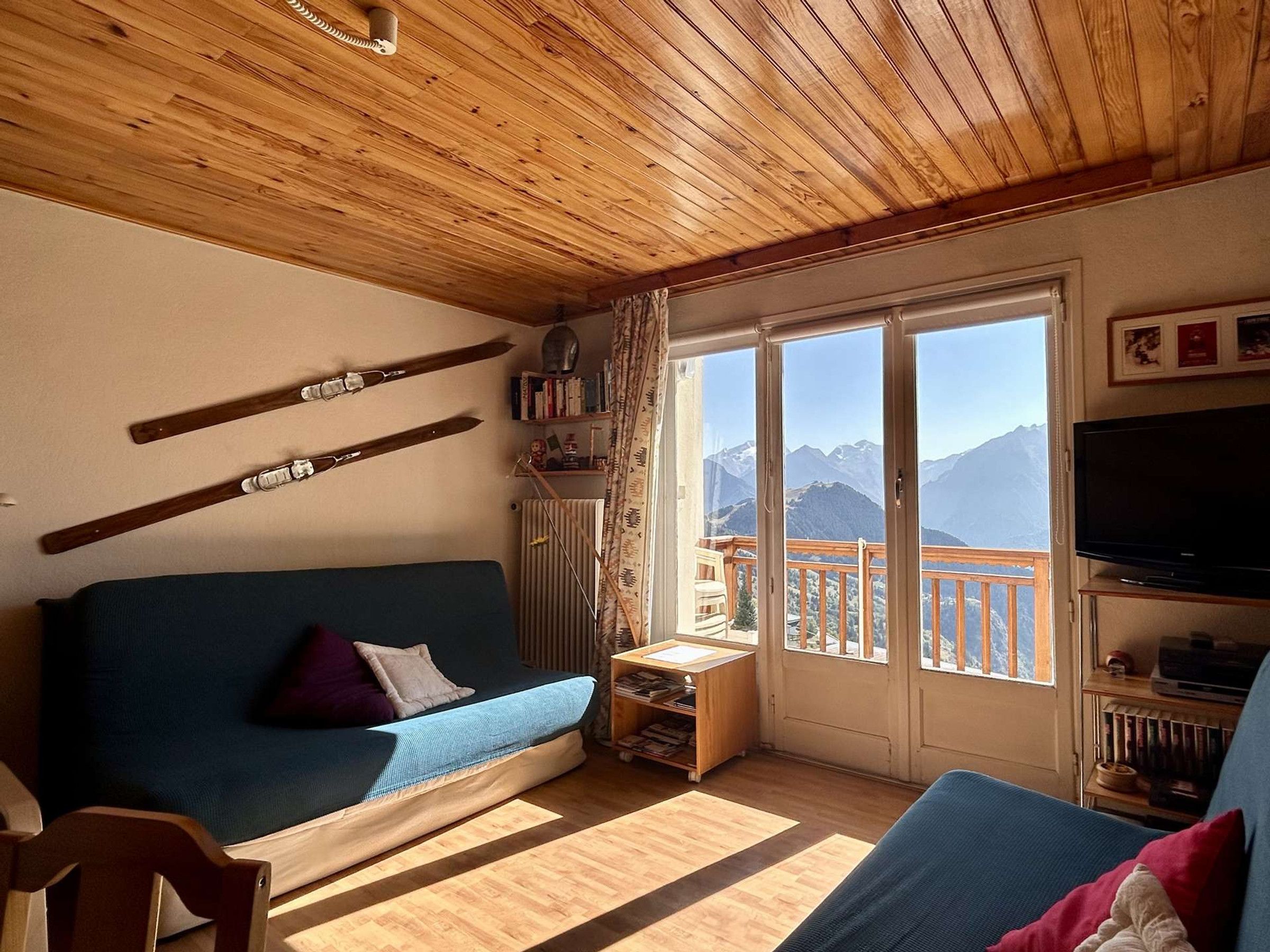 A cozy room with a wooden ceiling, a sofa, a TV, and a view of mountains through large windows.
