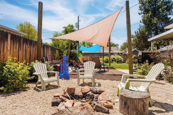 Un jardin avec un foyer, des fauteuils Adirondack, une clôture en bois et une bâche bleue servant de pare-soleil.