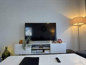 A modern living room with a flat-screen TV mounted on a white entertainment unit, a floor lamp, and a small plant. - Peaceful apartment in the village center - 5 guests (Villard-de-Lans)