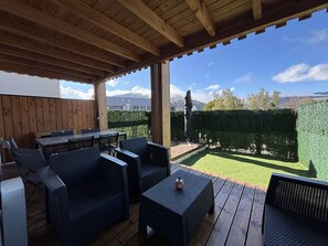An outdoor patio with wooden flooring, wicker furniture, and a view of a neatly trimmed hedge and distant mountains. - Peaceful apartment in the village center - 5 guests (Villard-de-Lans)