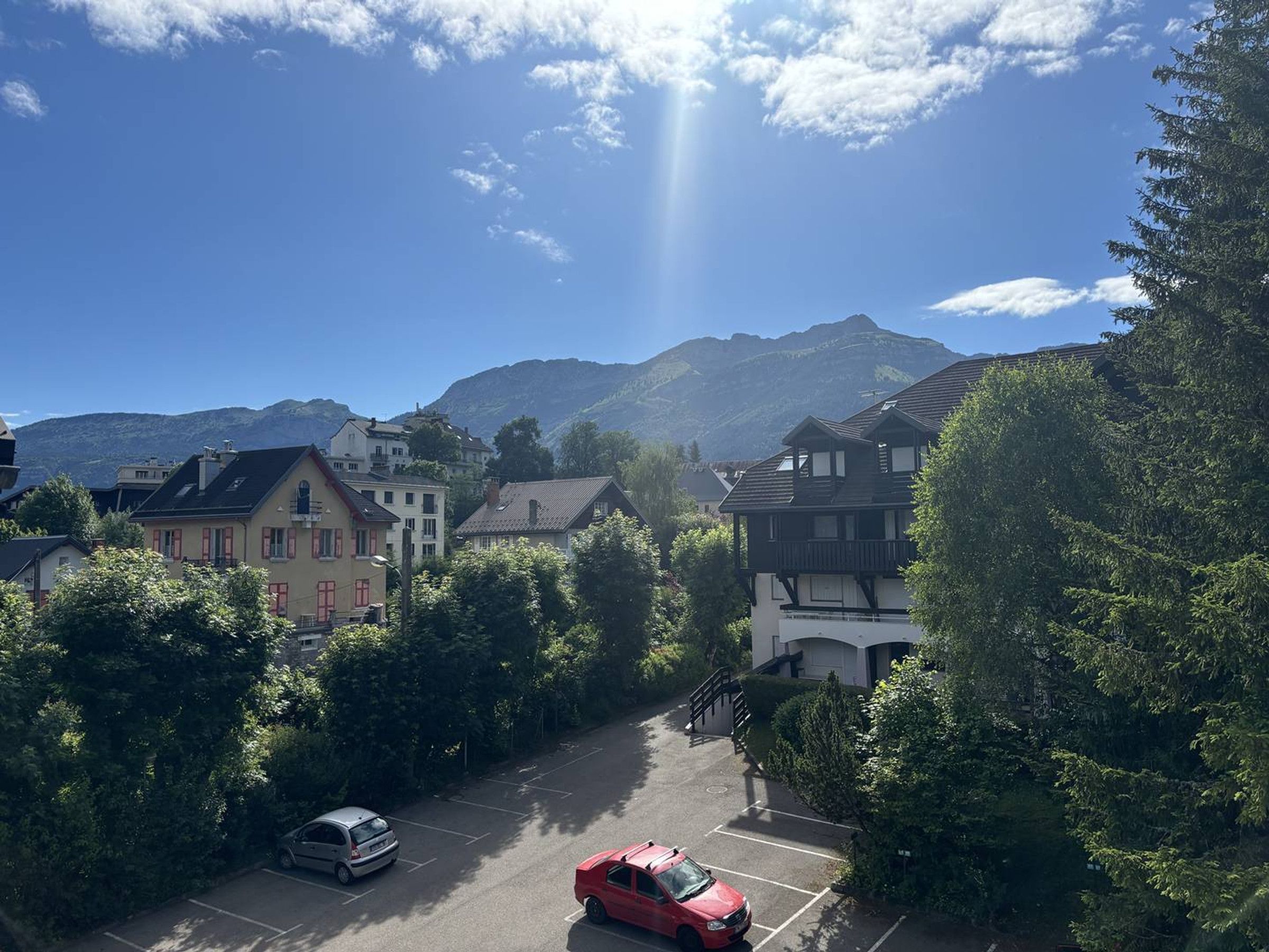 Ein Parkplatz mit einem roten Auto, umgeben von Bäumen und Gebäuden, mit Bergen im Hintergrund.