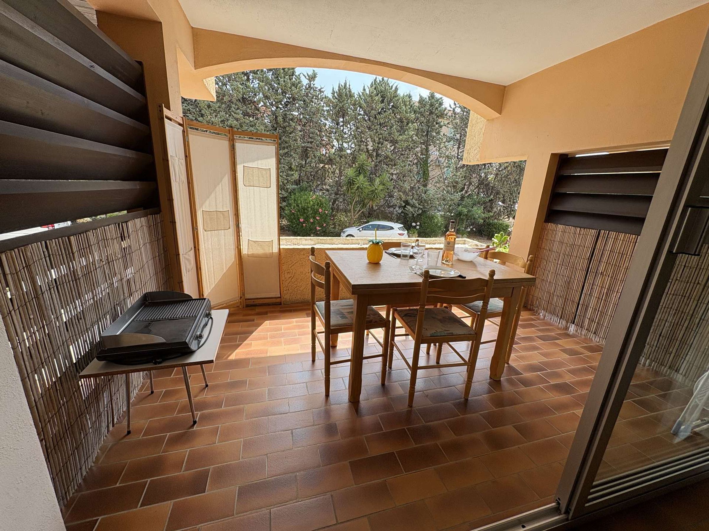 Un balcon avec une table et des chaises, un barbecue et une vue sur les arbres et une voiture.