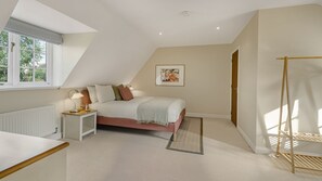 A bedroom with a sloped ceiling, a bed, a bedside table with a lamp, a radiator, and a window with a view of trees. - Rowan House - Bourton-on-the-Water, Sleeps 7 (Bourton-on-the-Water)