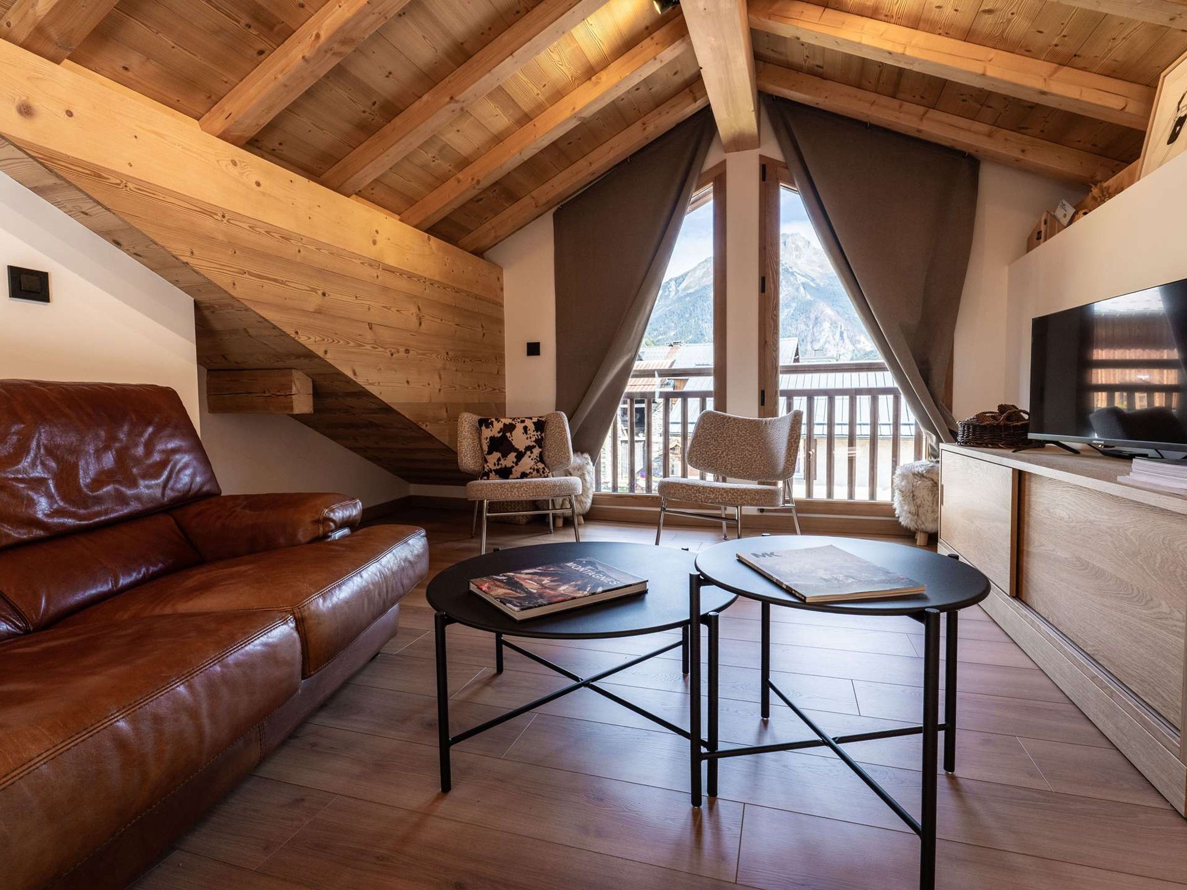 Una habitación acogedora con techo de madera, un sofá de cuero, una pequeña mesa con libros, una silla y un televisor sobre un mueble de madera.