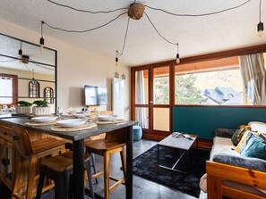 A dining area with a wooden table set for four, a television, and a window with a view of buildings. - Apartment mountain view - 4 guests (Saint-Lary-Soulan)