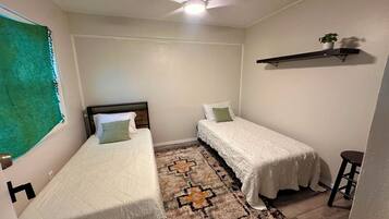 Une petite chambre propre, avec deux lits simples, un ventilateur de plafond, un tapis à motifs et une petite étagère sur laquelle est posée une plante.