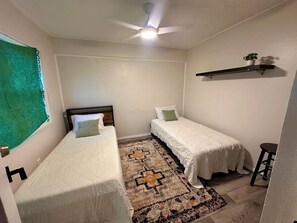 Une petite chambre propre, avec deux lits simples, un ventilateur de plafond, un tapis à motifs et une petite étagère sur laquelle est posée une plante.
