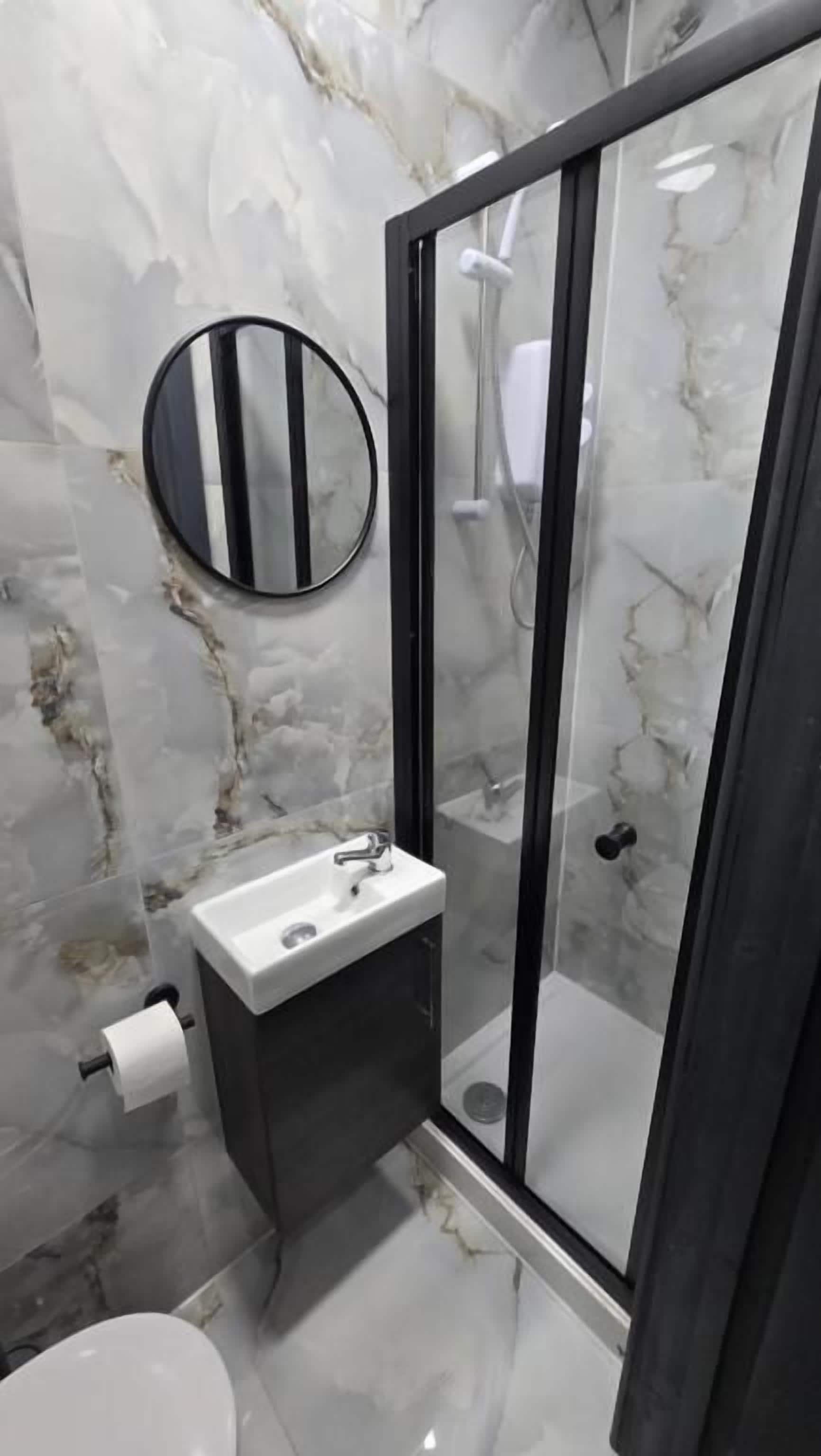 A bathroom with a marble wall, a round mirror, a sink, and a glass-enclosed shower.